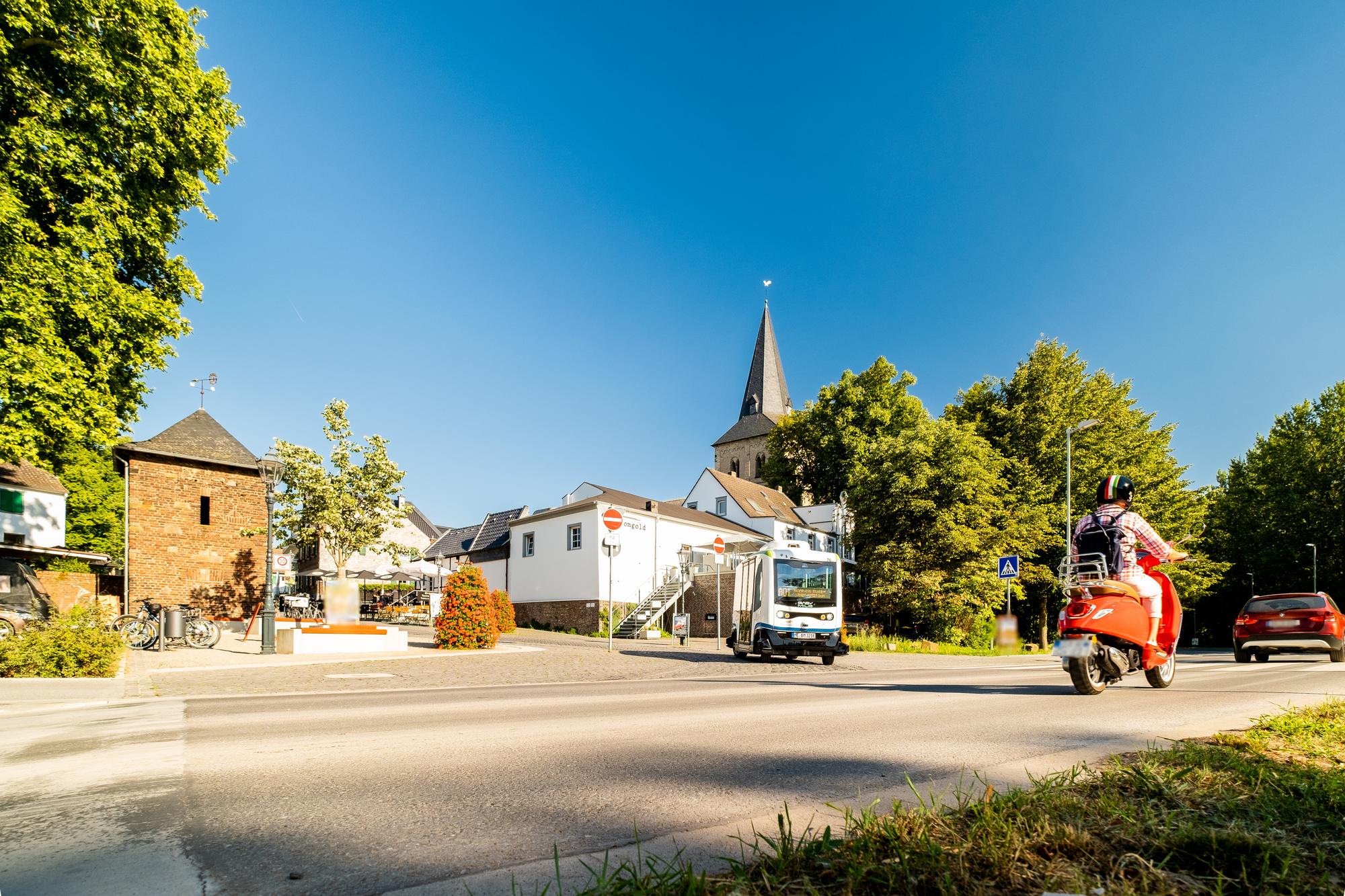 Vom Straßenrand aus blickt man auf die Straße, auf der ein Scooter vorbeifährt. Im Hintergrund biegt ein autonomer Kleinbus ab. 
