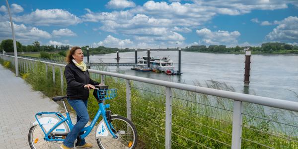 Eine Frau steht mit einem Stadtrad auf der Rheinpromenade und schaut auf den Rhein.
