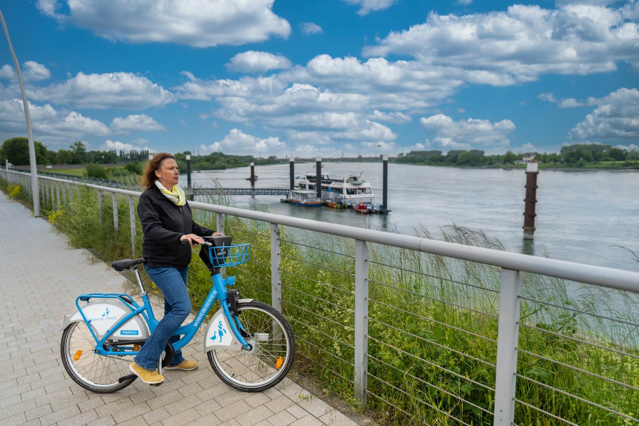 Eine Frau steht mit einem Stadtrad auf der Rheinpromenade und schaut auf den Rhein.